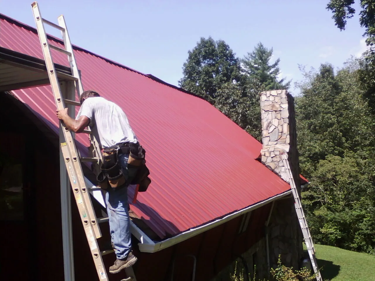 Roofer on a ladder beside a red metal roof and stone chimney — Soffit & Fascia Repair by Roofing Brothers in Alvaton